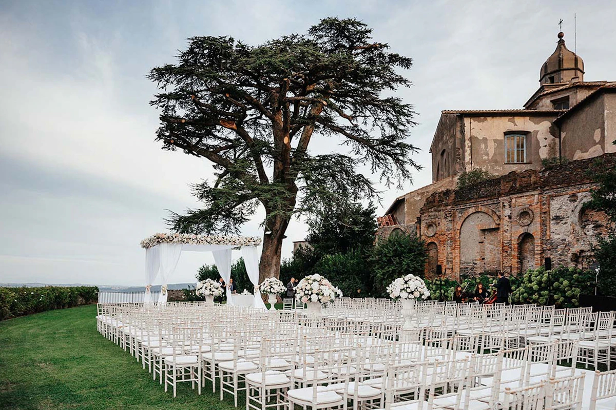Cérémonie en plein air sous un grand cèdre, chaises blanches face à une arche fleurie