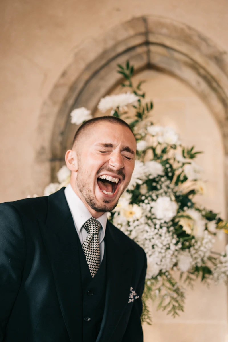 Marié qui éclate de rire, les yeux fermés, devant une arche de fleurs blanches