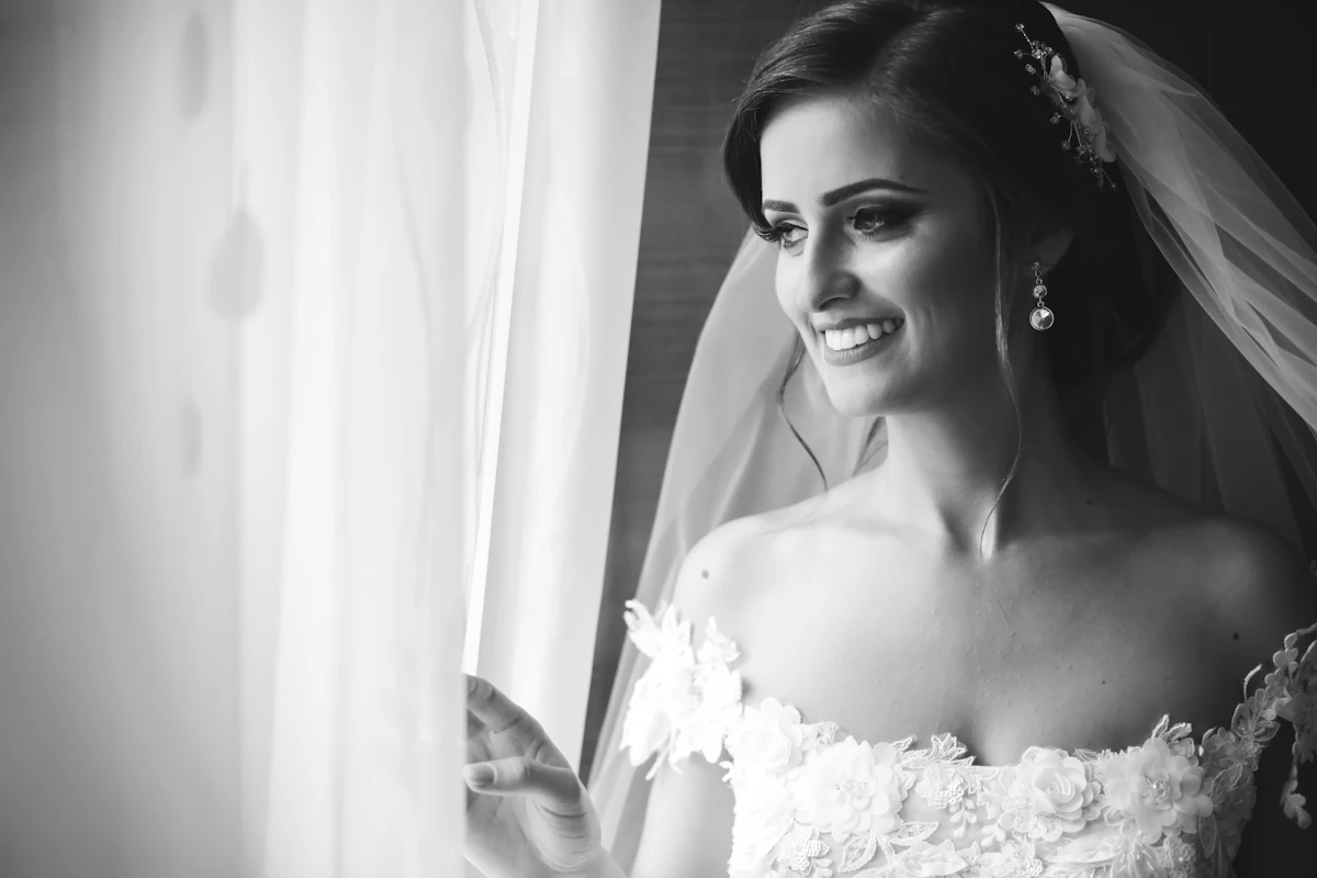 Mariée qui sourit près d’une fenêtre, voile sur les épaules, en noir et blanc