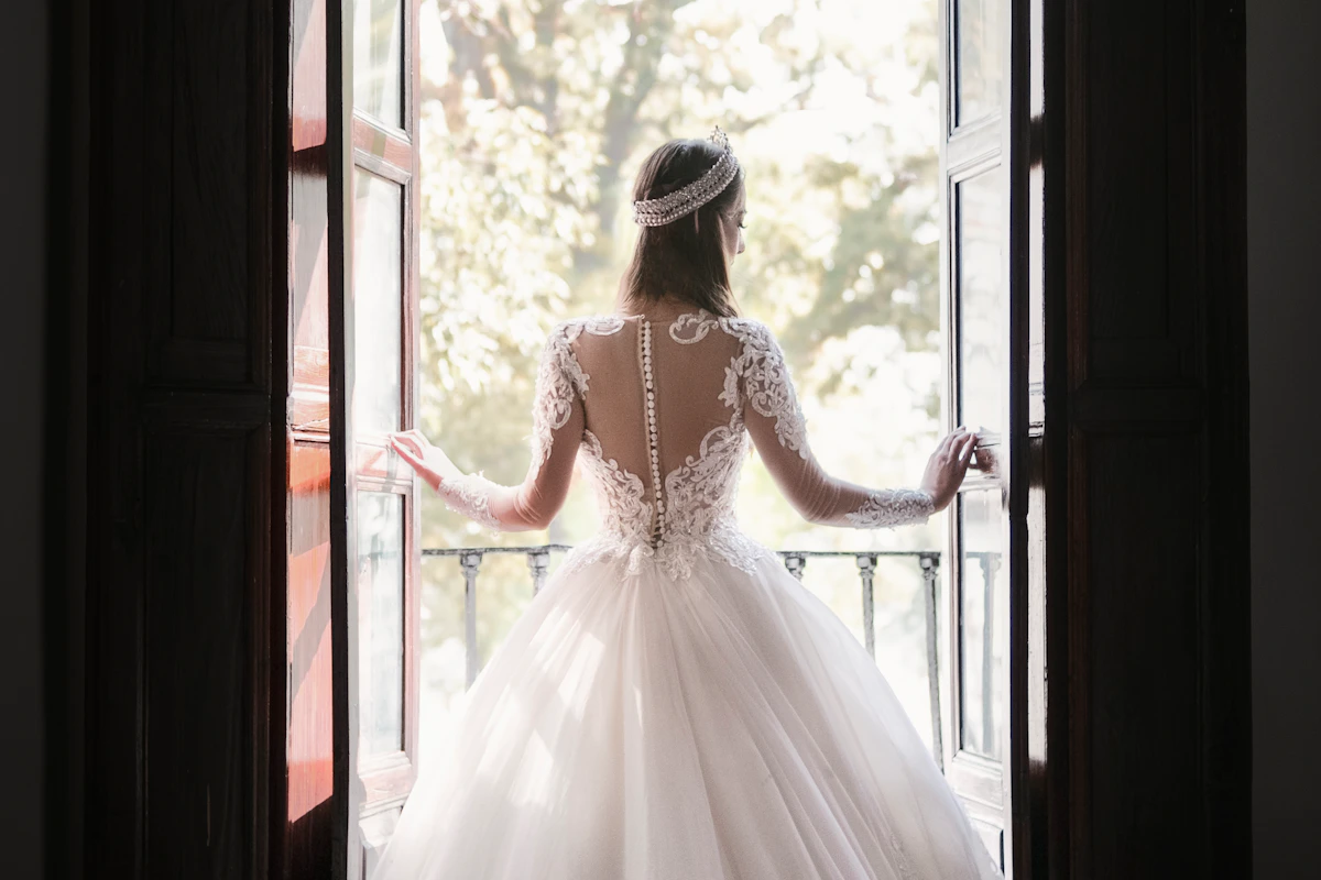 Mariée de dos devant une porte-fenêtre ouverte sur un balcon, robe en dentelle